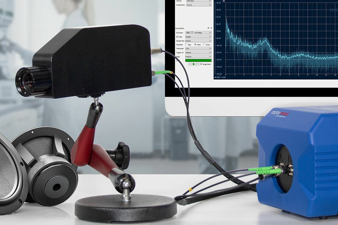 Single-point Laser Doppler Vibrometer with connected fiber head mounted on an adjustable stand, linked to a processing unit; a monitor in the background displays a frequency spectrum.